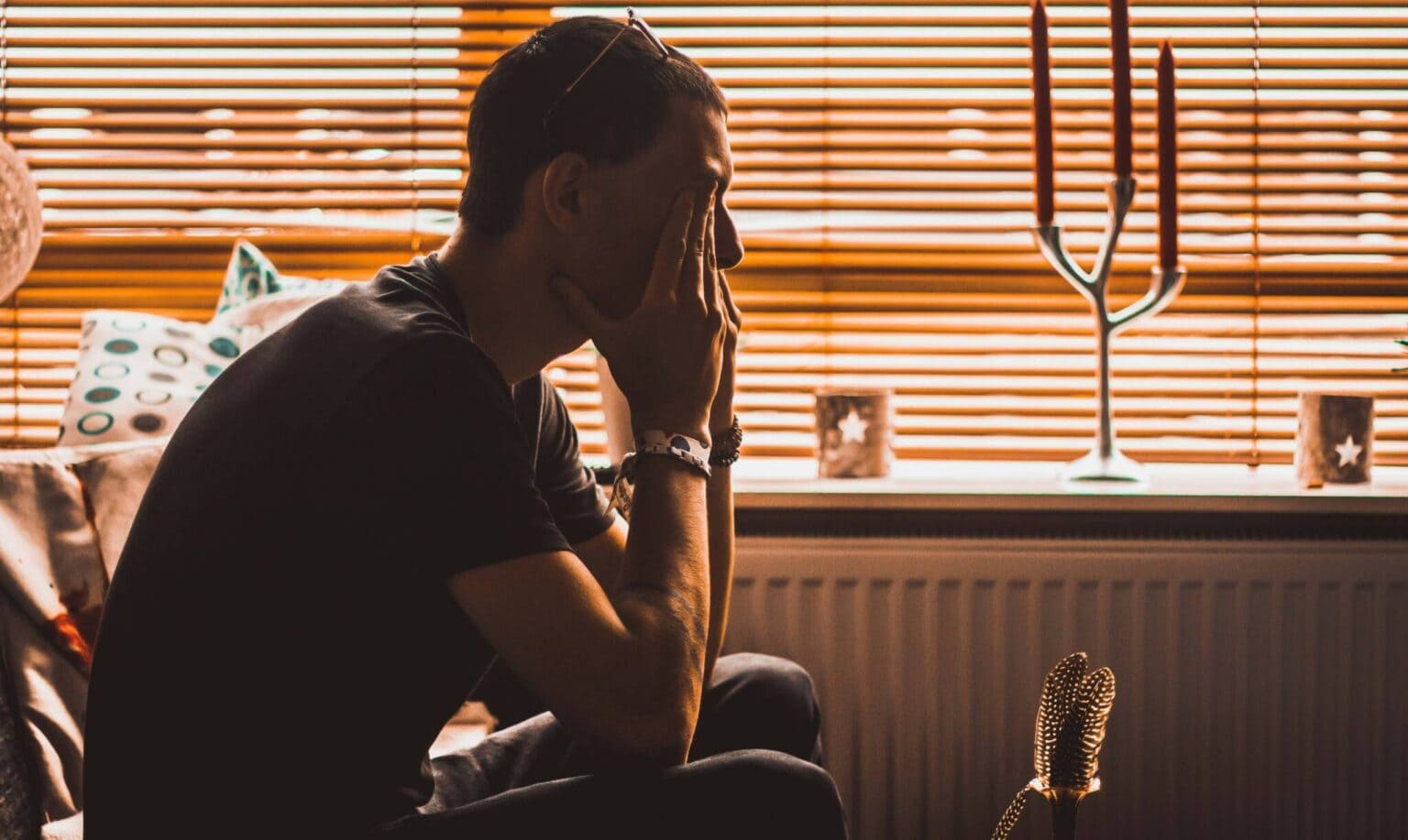 Depressed man sitting alone in a dimly lit room with blinds and candles.