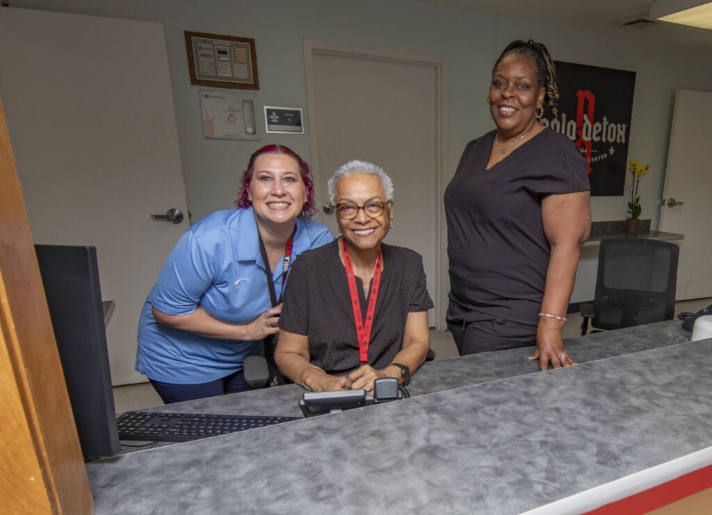 Three smiling women at NOLA Detox reception desk in New Orleans.