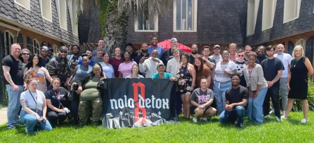 Group of alumni holding NOLA Detox banner outdoors in front of a building.